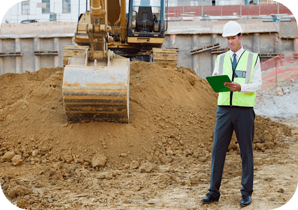 Man in high visibility vest and white hard hat writing on a clipboard with a yellow construction vehicle on a dirt mound behind him.