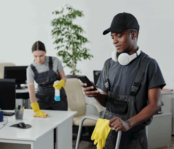 Young male cleaner is on the cleaning service business software whilst his female colleague cleans the office desk