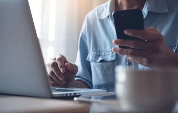 Woman in a blue shirt using their phone whilst they are on the laptop.