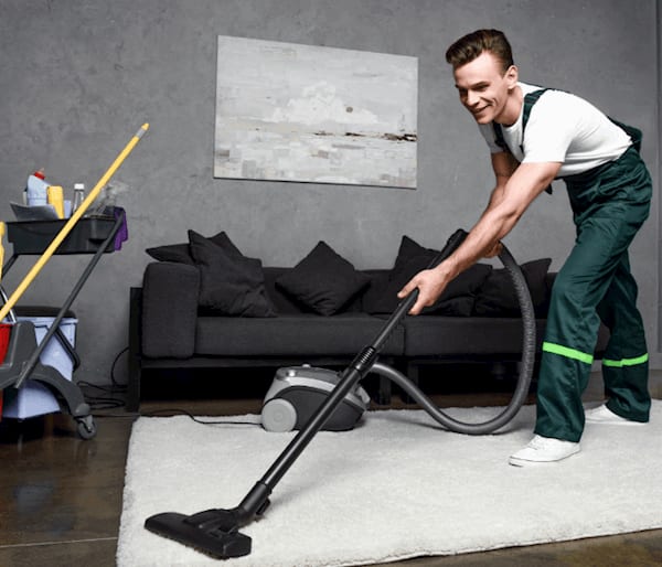 Man in white T-Shirt and green overalls is vaccuming a rug in a house