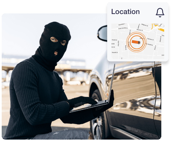 A car thief wearing all black, gloves and a ski mask is on a laptop outside of a vehicle. In the top right of the image there is a location notification with the tracked vehicle on the live map which has multiple orange circles around it