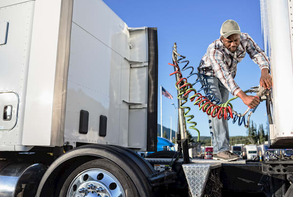 A trucker in a baseball cap and a flannel shirt is fixing his air brakes on a truck being tracked by fleet management software.
